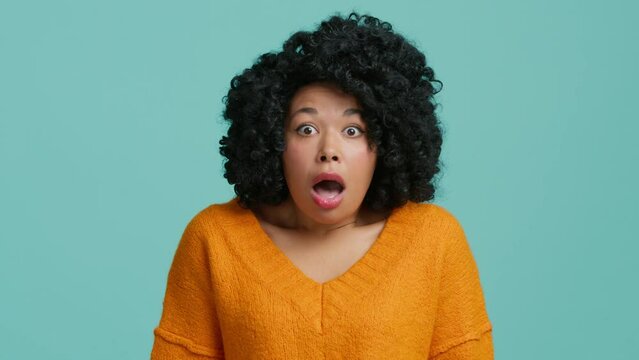 Close up portrait of shocked amazed scared African American woman with wide open mouth touching face in astonishment and wonder. Curly hair fancy model slow motion studio shot on blue background 4K