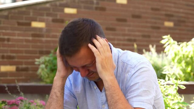 Mature man in a blue shirt rubs his forehead tiredly, suffering a headache, sitting hunched on a wooden bench among green bushes in a city park