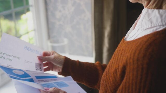 Senior woman standing by window at home opening UK energy bill during cost of living crisis looking worried - shot in slow motion