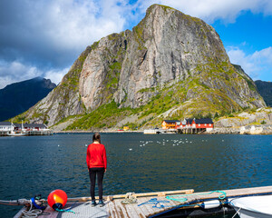 a girl in a red sweatshirt stands in the harbour looking up at a mighty mountain in the reine village in the lofotenland in norway, the mighty Norwegian fjords and picturesque fishermen's villages