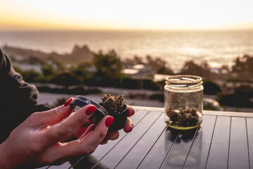 Hands with red nails putting a weed bud in a marijuana grinder, glass jar with marijuana buds and water pipe (bong) with beautiful sunset sky and sea - obrazy, fototapety, plakaty