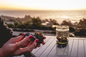 Fotobehang Hands with red nails putting a weed bud in a marijuana grinder, glass jar with marijuana buds and water pipe (bong) with beautiful sunset sky and sea © Samuel Ponce