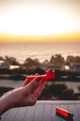 Hand with red nails holding a orange plastic marijuana pipe with weed and orange lighter with beautiful sunset sky and sea