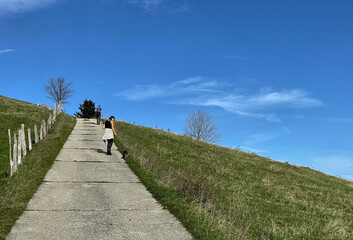 Wanderer mit Hund gehen auf einem betonierten Weg neben einem Holzzaun vor strahlend blauem Himmel bergauf