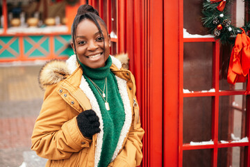 African american woman standing street outside near Christmas Tree fair market, red telephone box. New Year atmosphere, winter vacation, cold weather
