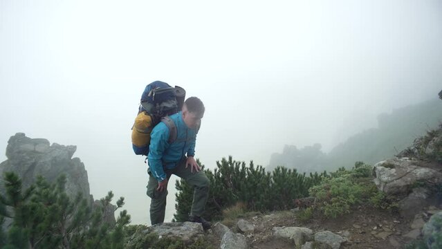 A man with a backpack climbs rocky mountains into the fog. A tourist conquers a mountain peak in the rain. A boy walks uphill next to large stones in the Carpathians. Concept: submission, fatigue. 4k