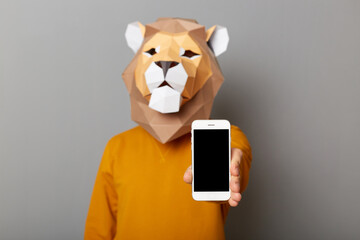 Portrait of unknown man wearing lion mask and orange jumper showing smart phone with empty black...