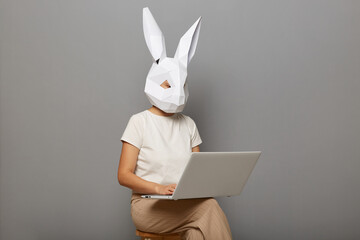 Image of unknown female wearing bunny mask and white t-shirt isolated over gray background, sitting on chair and working on personal computer, doing her work. - obrazy, fototapety, plakaty