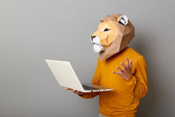 Portrait of angry anonymous man wearing lion mask and orange sweatshirt standing with laptop in hands, working online, having problems with work, posing isolated over gray background.