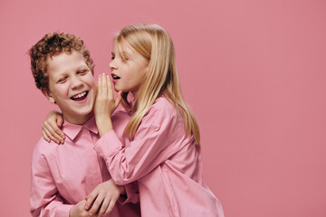 cute, beautiful, happy children are standing in pink clothes on a pink background and the girl tells the boy secrets in his ear. Topics of friendship and relationships