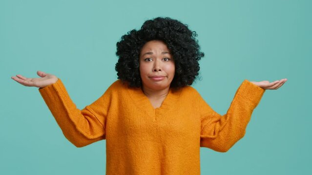 Dont know, who cares. Confused lovely beautiful African American girl shrugging shoulders, looking with uncertain clueless expression, demonstrating indifference doubts. Indoor studio blue background