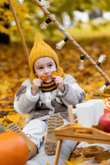 Cute little baby boy in a yellow knitted cap on a fall picnic. Cozy atmosphere, autumn vibe. Pumpkin, plaid, orange maple leaves as decor
