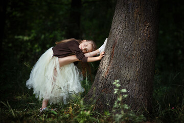 Pretty little slim girl doing stretching exercises as ballet dancer ballerina near big tree in summer forest or park, with leg raised up, dressed in fluffy dress and ballet shoes, dark background © upparadox
