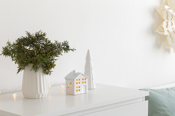 Christmas cozy winter home decor. New year interior decorations. Green fir branch in vase, decorative ceramic house and christmas tree, glowing garland lights. Stylish composition on the dresser.