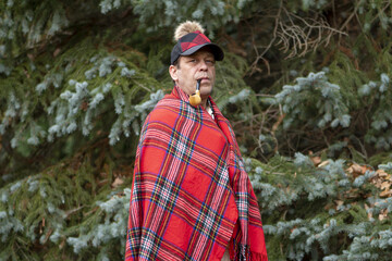 Portrait of an elderly man 45-50 years old with a smoking pipe in his mouth, with a plaid on his shoulders and a stylish cap against the background of fir trees.