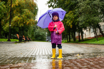 Child playing in autumn rain. Kid with umbrella. Outdoor fun for kids by any weather. Rain waterproof wear, boots and jacket for children