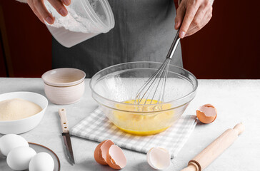 Woman pour flour into a bowl of fresh beaten eggs and mix with whisk on a gray table in the kitchen. Authentic female hands making a dough for homemade pie, Home baking concept, pie crust recipe