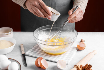 Woman pour milk into a bowl of beaten eggs and flour, mix with whisk on a gray table in the...