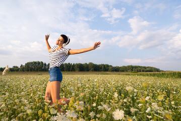 Fototapeta premium Young smiling woman wearing headphones listening music from her smartphone on the spring meadow full of white flowers