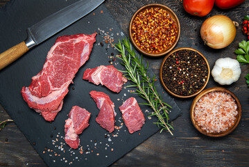 Sliced fresh meat on a black, stone board. Salt, pepper, herbs and spices with vegetables on a dark table. View from above.