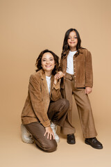 Fototapeta premium smiling mother and child in suede jackets and trendy clothes holding hands on beige background