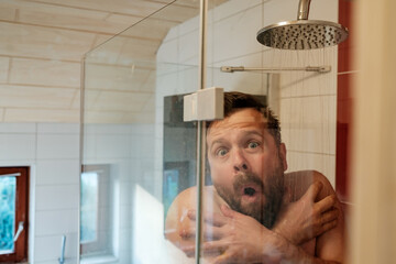 Man in the shower under cold water, freezes and hugs himself. Concept of savings and energy crisis.