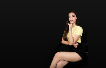 Young Asian girl with long dark hair touching her finger to her chin with second hand supporting her elbow sits on chair sideways with her legs crossed, wearing yellow t-shirt on black background 