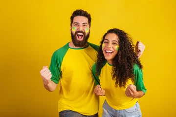 Fotobehang couple of brazil soccer supporters, dressed in the colors of the nation, black woman, caucasian man. Twisting and vibrating. © StockImageBrasil