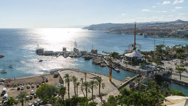 Aerial Time lapse of people walking at eilat boardwalk hotel in front of the red sea, the hotels cityscape and moab mountains at the background, Eilat, Israel. zoom effect.