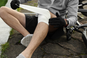 Fototapeta na wymiar Man applying bandage onto his knee near bicycle outdoors, closeup