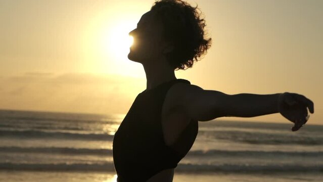 Silhouette of a young woman. She stands sideways to the camera, a huge disk of the sun shining behind her. The woman smoothly raises her hands and spreads them freely to the sides.