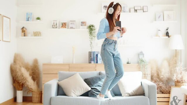 On the sofa in her living room, a young Asian woman dances. Asian woman with a smile relaxing at home, happy and in good mental and physical health, wearing headset and listening to music