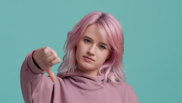 Young hipster woman with pink hair in hoodie sweater making good-bad sign isolated on teal blue background. Girl make choice, or makes a decision thumb up or thumb down, like or dislike, yes or no 4K