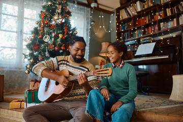 Happy black father playing acoustic guitar to his daughter on Christmas at home.