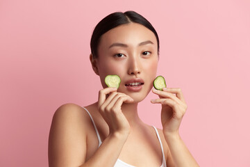 Asian Woman Holding Cucumber. Portrait of Beautiful Girl Hold Slice of Cucumber. Smiling Young Woman Looking at Camera. Concept of Skincare. Isolated on Pink Background. Studio Shot 