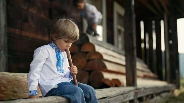 Little boy playing on woodwind wooden flute - ukrainian sopilka. Folk music concept. Musical instrument. Kid in traditional embroidered shirt - Vyshyvanka. Child sitting on porch of old wooden house