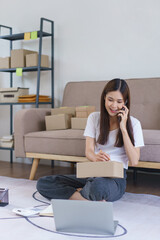 Online business concept, Asian business woman talks on smartphone and writes address on parcel box