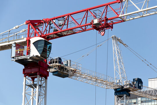 A close-up of two large, modern construction cranes, one red and one blue, are seen on a clear, blue sky day.