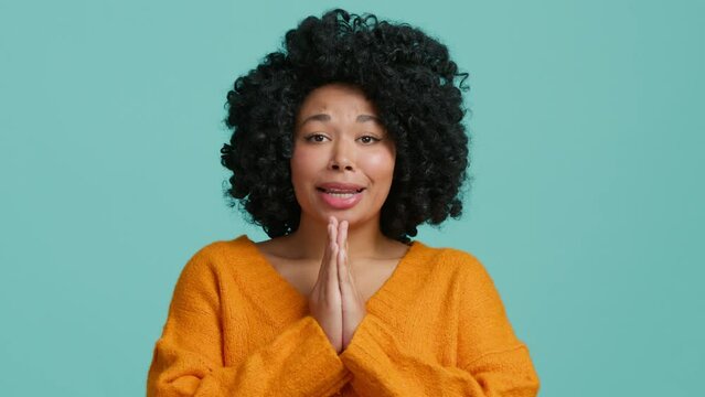 Young African American woman 20s in orange sweater look camera, holding hands folded in prayer, beg about something good, asking for something on blue background in studio. People lifestyle concept 4K