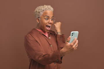 Fototapeta premium Young amazed African American woman shouting and making YES gesture looking at screen of mobile phone rejoicing at victory of sports team or jackpot in online casino, stands on brown background