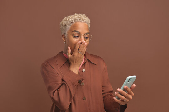 Young stunned optimistic African American woman in casual clothes covers mouth with hand after reading news on phone with gossip from life of celebrity stands in brown studio. WoW emotion