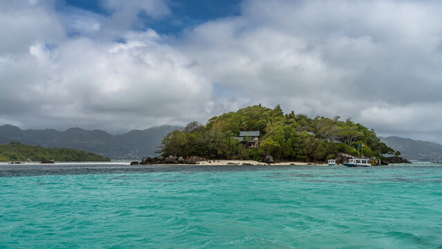 The secluded island is overgrown with tropical vegetation. The roofs of the houses are visible among the trees. The boat is moored at the sandy beach. Turquoise water. White clouds in a blue sky.