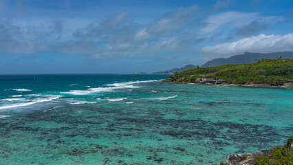 Fototapeta na wymiar Beautiful tropical seascape. Waves of turquoise ocean foam over coral reefs. On the shore, overgrown with tropical vegetation, the houses of the hotel are visible. Blue sky with clouds. Seychelles