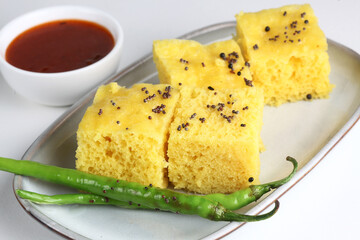 This is a vegetarian culinary dish called Dhokla served in a white plate with chutney in a white bowl on a white background