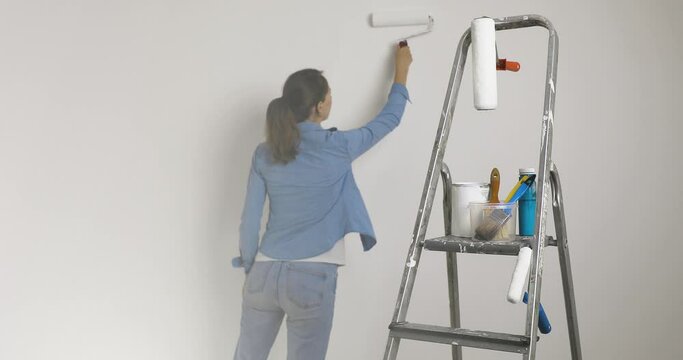 	
Ladder with paint brushes, buckets and tools a woman painting a wall in background. Home improvement.