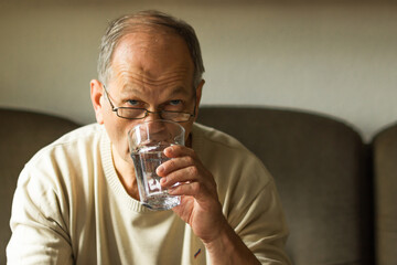 Caucasian senior man in white sweater take capsule and drinking clear water from glass on sofa at home, feels like sick.