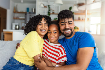 Attractive happy family hugging attractive mother handsome father and adorable small daughter sitting together on couch in living room at home smiling looking at camera