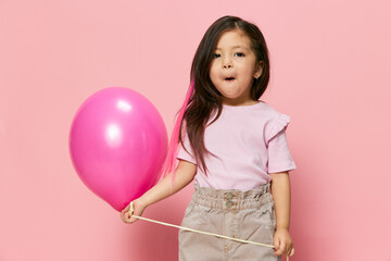 Obraz na płótnie Canvas happy, joyful, playful little girl playing with a pink air balloon standing in stylish clothes on a pink background with an empty space for an advertising mockup. Themes of holidays, happiness