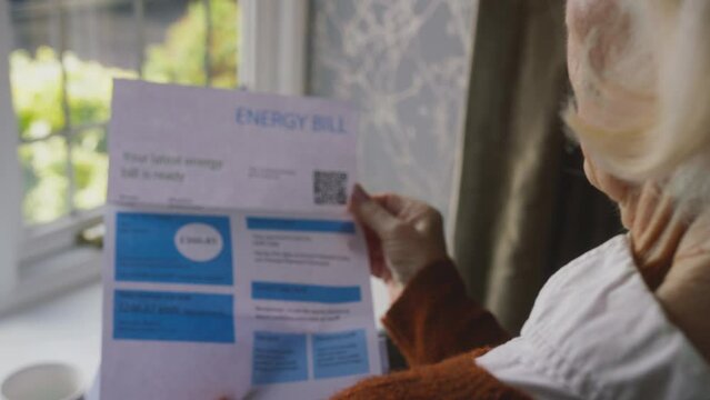 Senior woman standing by window at home with UK energy bill during cost of living crisis looking worried - shot in slow motion