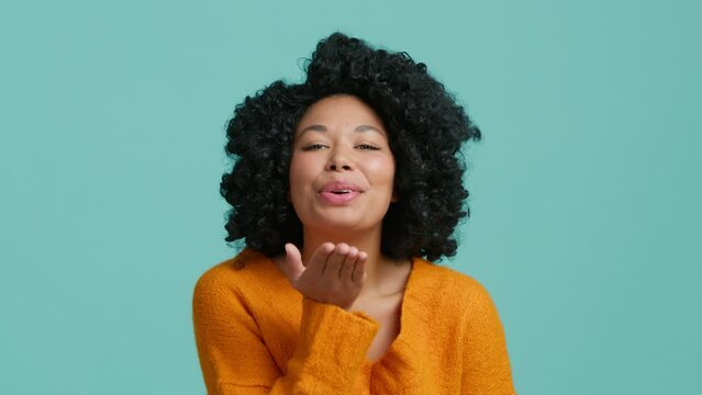 Bye gesture with air kiss by cute beautiful African American girl on pastel blue isolated studio background. Happy lady with curly hair send kiss you blowing from palm of the black skinned hand slowmo